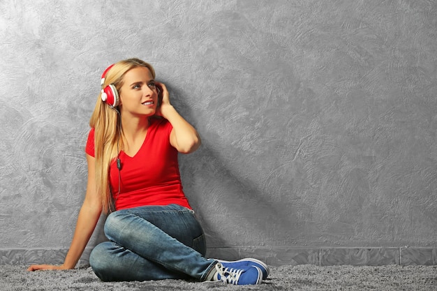 Young woman sitting on the floor and listening to music on a grey wall background