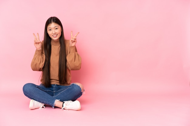 Young woman sitting on the floor on pink showing victory sign with both hands