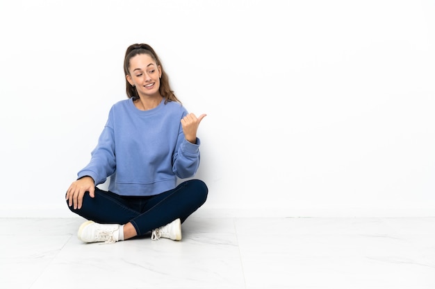 Young woman sitting on the floor pointing to the side to present a product