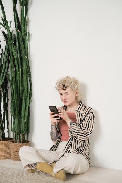 Young woman sitting on the floor in the room with plants and reading a book online using her mobile phone