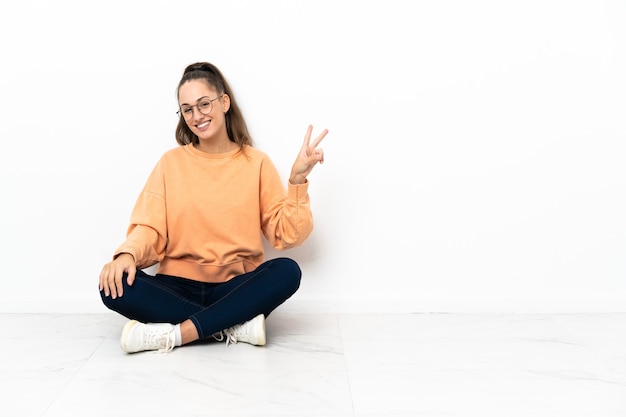 Photo young woman sitting on the floor smiling and showing victory sign