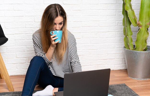 Giovane donna seduta sul pavimento con il suo portatile in possesso di una tazza di caffè