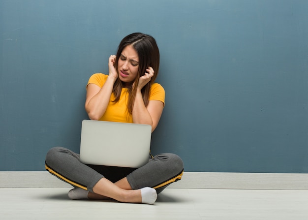 Young woman sitting on the floor with a laptop covering ears with hands