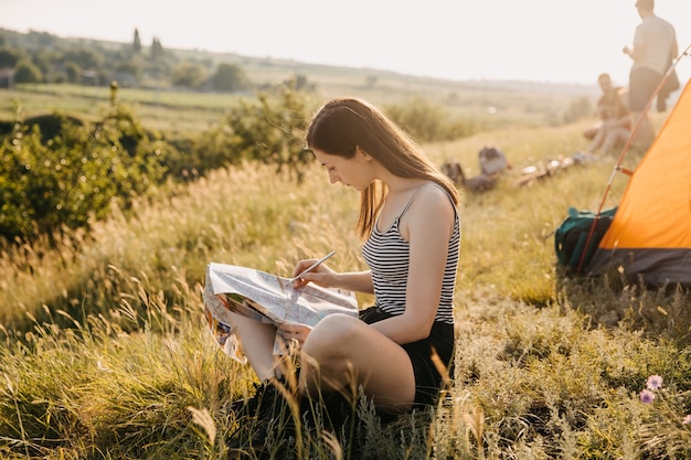 Young woman sitting on grass by the tent, holding a map.