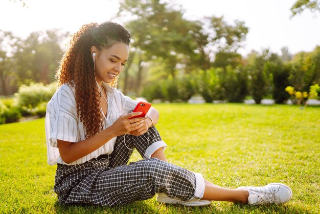 Young woman sitting on green grass in park with phone Pretty woman listening to music