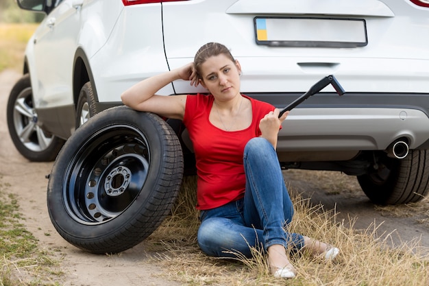 壊れた車で地面に座って、ホイールレンチを見ている若い女性