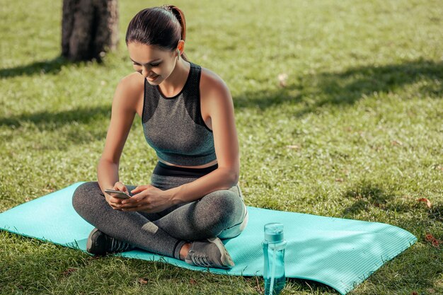 Young woman sitting on mat and texting on smartphone in the park