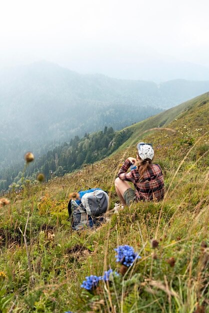 Foto giovane donna seduta su un prato in una valle di montagna accanto a uno zaino che beve acqua in un paesaggio di escursioni