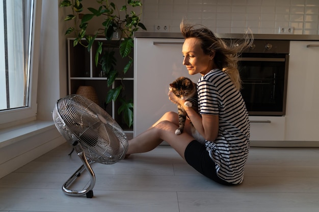 扇風機の近くに座っている若い女性は、猫が夏の暑さと息苦しさから涼むのを助けます
