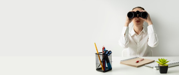 Young woman sitting in the office at the workplace looking through binoculars Banner