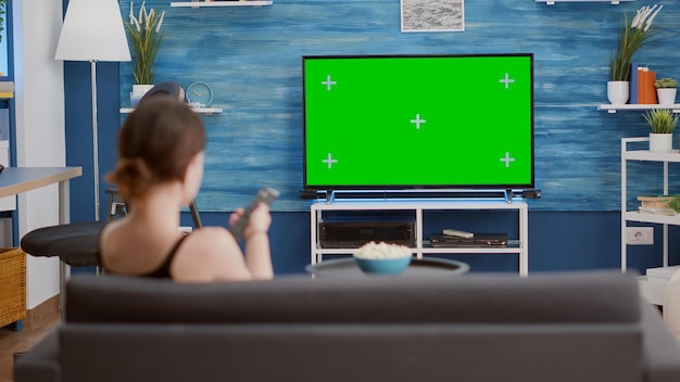 ソファに座ってテレビの緑色の画面を見て、モダンなリビングルームでチャンネルを切り替える若い女性。クロマキーディスプレイのテレビリモートザッピングプログラムを使用してリラックスしている女の子の背面図。