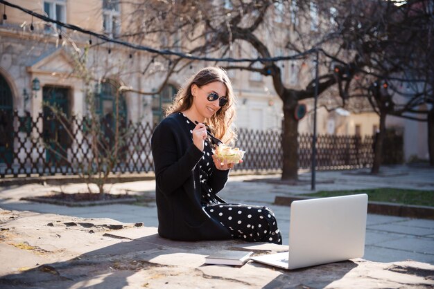 Young woman sitting outside eating vegetable salad and working\
at laptop spring sunny day outdoor work lunch