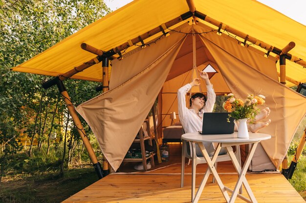 Young woman sitting outside tent and drinking coffee in forest Woman relaxing and looking at tree covered forest Happy woman admiring nature while camping on Ukrainian nature