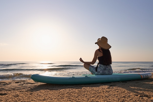 Giovane donna seduta su una tavola da paddle con una postura rilassante all'alba sulla spiaggia