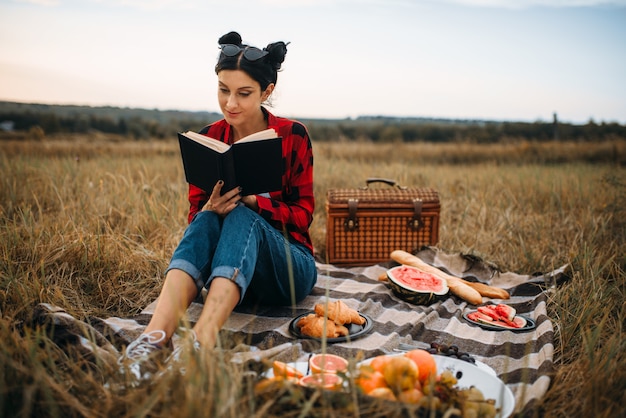 Молодая женщина сидит на пледе и читает книгу, пикник в летнем поле. Романтический пир, с праздником