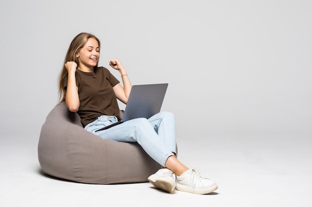 Young woman sitting on pufff with laptop with win gesture isolated on white wall