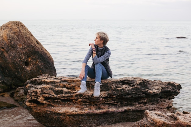 Young woman sitting on the rock near sea in autumn or summer time