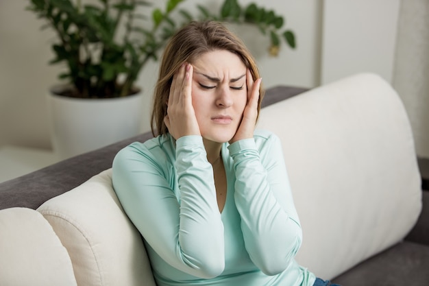 Молодая женщина сидит на диване и страдает от головной боли