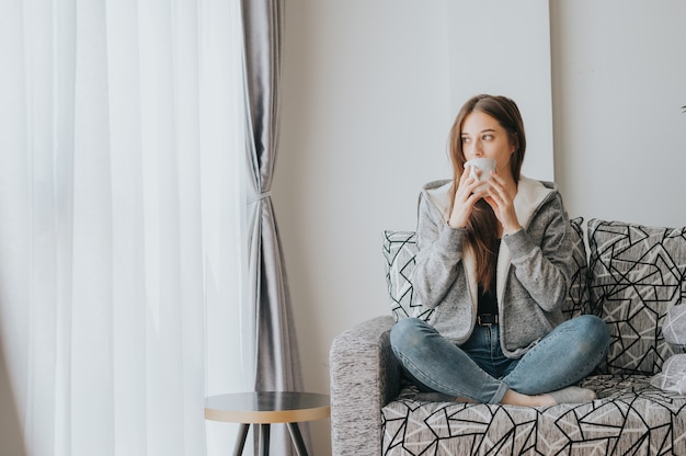 Giovane donna seduta sul divano che indossa un maglione sorseggiando caffè o tè dalla tazza bianca al mattino durante la stagione invernale guardando fuori