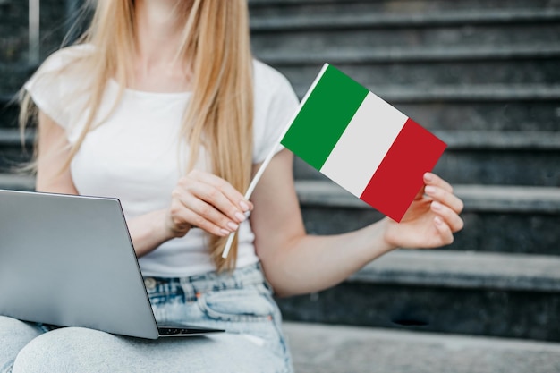 Foto giovane donna seduta sulle scale con un computer portatile e con in mano la bandiera dell'italia