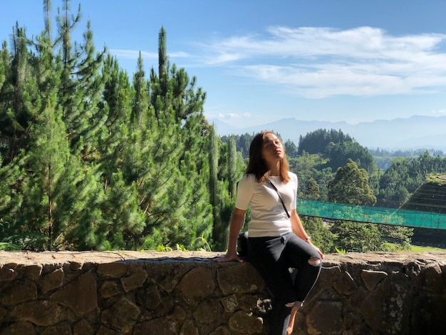 Young woman sitting on stone wall against trees