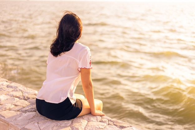 Young woman sitting on stone wall enjoying herself watching sunset at the beach