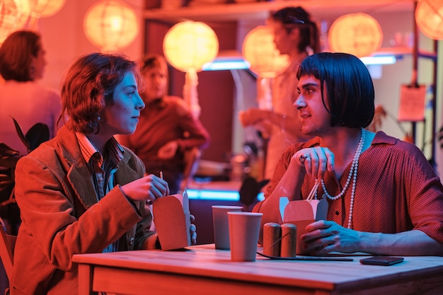 Молодая женщина сидит за столом и ест еду вместе со своим другом-геем в баре