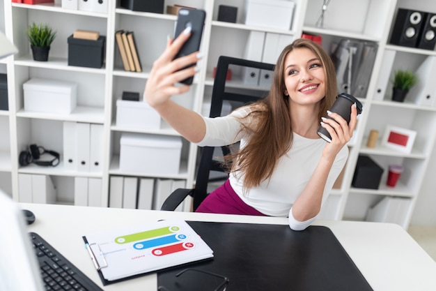 A young woman sitting at a table in the office and holding a glass of coffee and a phone.