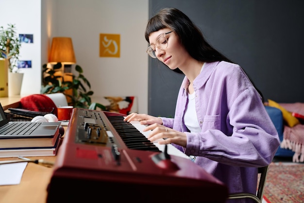 Молодая женщина сидит за столом и играет песню на синтезаторе в своей комнате