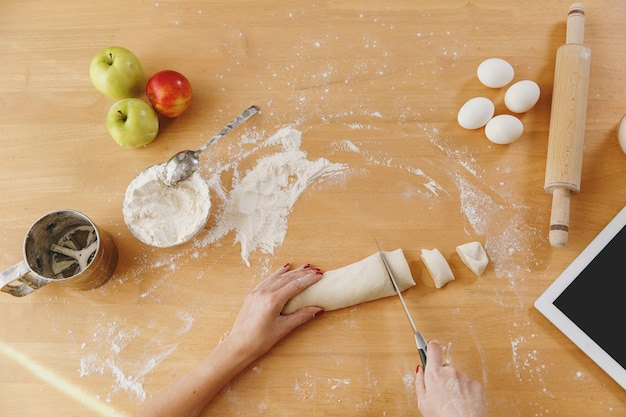 A young woman sitting at a table with tablet, cuts a dough with a knife into pieces at home in the kitchen. Cooking home. Prepare food. Top view.