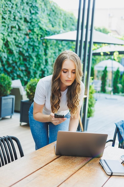 Young woman sitting on the terrace of a cafe on the street of a modern cafe Woman sitting at table with phone and using laptop outdoors Remote work in a summer cafe
