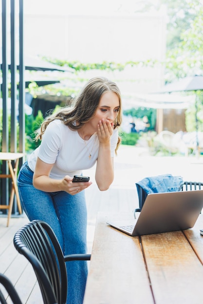 Молодая женщина сидит на террасе кафе на улице современного кафе Женщина сидит за столом с телефоном и использует ноутбук на открытом воздухе Удаленная работа в летнем кафе
