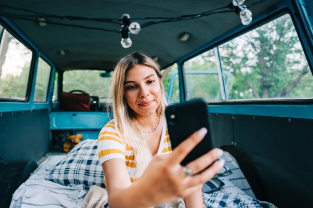 Young woman sitting in a van and using mobile phone, looking on screen, outdoors in nature. enjoying summer, travel concept