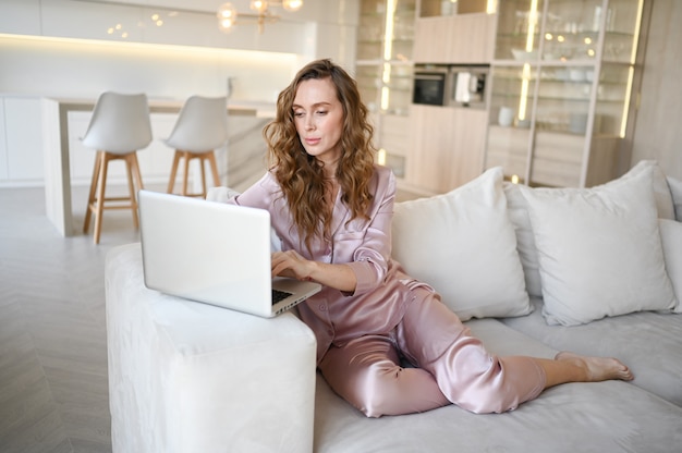 Young woman sitting on a white sofa in scandinavian style living room kitchen interior and working on a laptop.