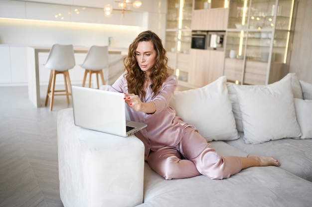 Young woman sitting on a white sofa in scandinavian style living room kitchen interior and working on a laptop.