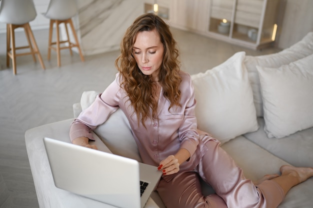 Young woman sitting on a white sofa in scandinavian style living room kitchen interior and working on a laptop.