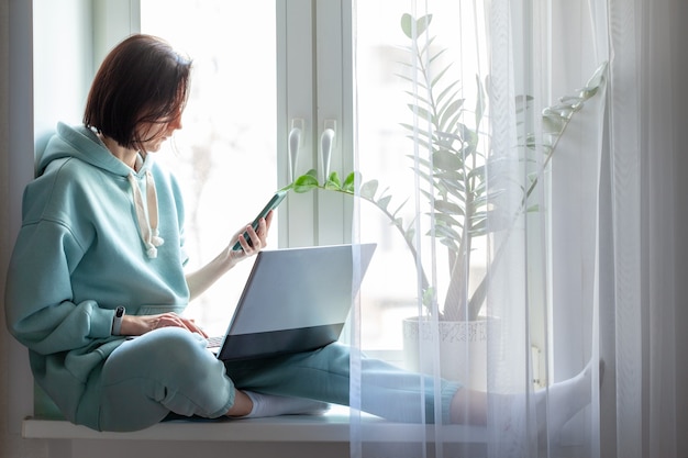 Young woman sitting on a windowsill with notebook and mobile phone. Girl working at home, studying on laptop. Concept freelance