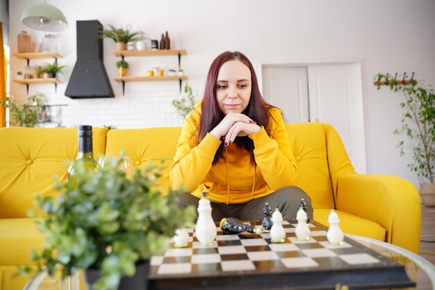 Young woman sitting on yellow sofa and playing chess in room Female playing in logical board game with herself