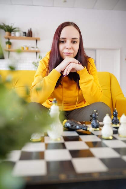 Young woman sitting on yellow sofa and playing chess in room Female playing in logical board game with herself