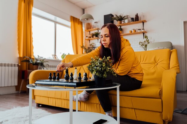 Young woman sitting on yellow sofa and playing chess in room Female playing in logical board game with herself