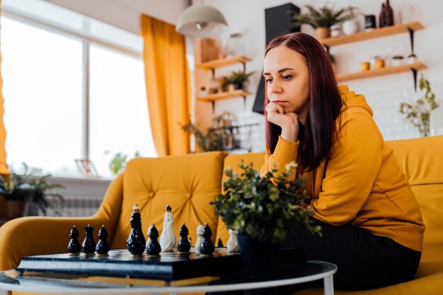 Young woman sitting on yellow sofa and playing chess in room Female playing in logical board game with herself