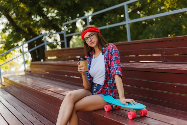 세련된 옷을 입고 젊은 여자 스케이팅 skatepark에서 스탠드에 앉아 밝고 화창한 날에 커피를 마신다