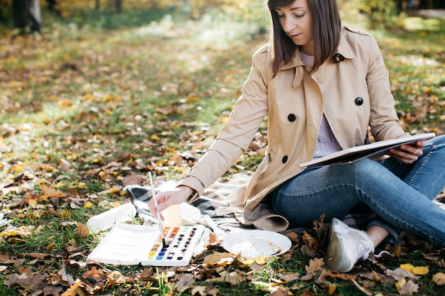 Молодая женщина делает зарисовки возле озера в осеннем лесу.