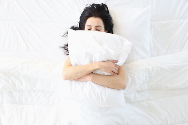 Young woman sleeping in white bed with pillow on her face top view. Comfortable and soft bedding concept