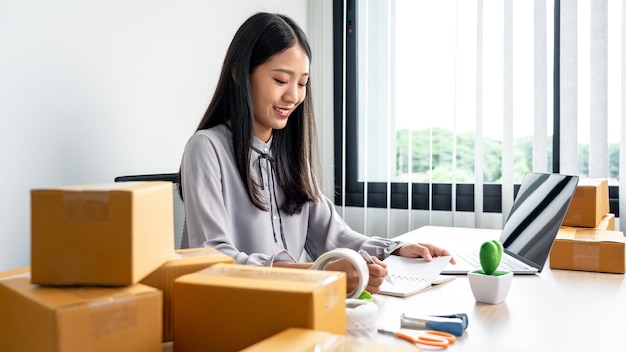 自宅で働く若い女性の中小企業の起業家は、オンライン顧客からの注文を受け取り、製品を梱包する準備をしながらデータを確認し、クライアントに送信します