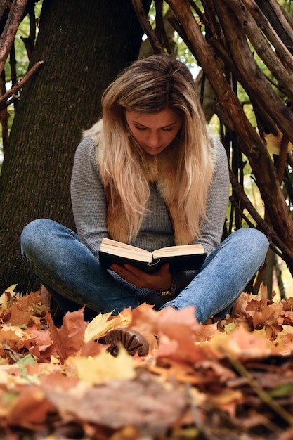 A young woman under a small hut sits on yellow leaves and reads a book