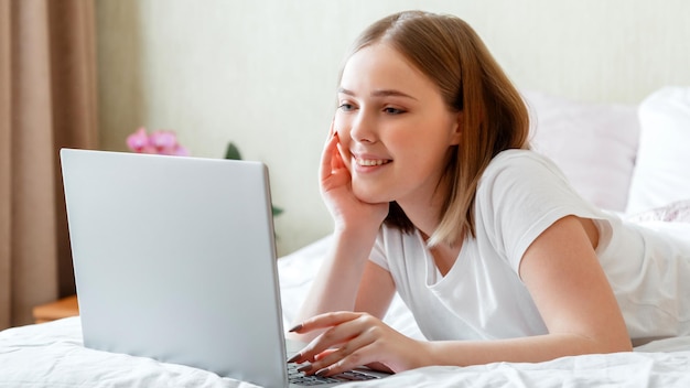 Молодая женщина улыбается и работает с ноутбуком, лежа в постели утром дома Счастливая девушка в пижаме учится онлайн или планирует свой день, использует ноутбук утром в спальне Длинный веб-баннер