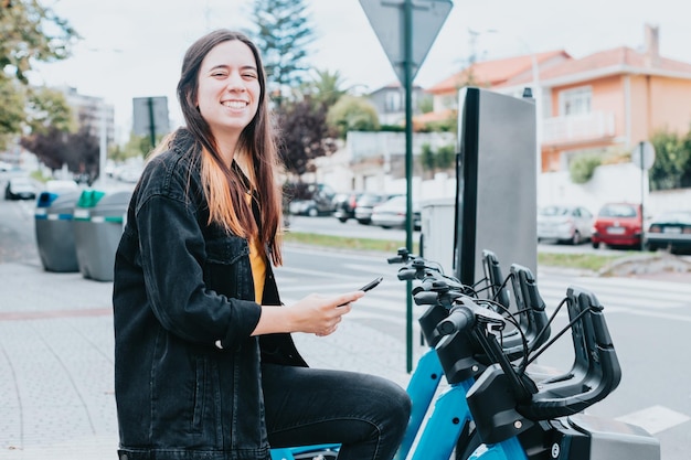 自転車のロックを解除してレンタルするために携帯電話をチェックする笑顔の若い女性 利用可能な自転車とバッテリーの状態を確認する 電気輸送の概念 再生可能な資源 街中を移動する