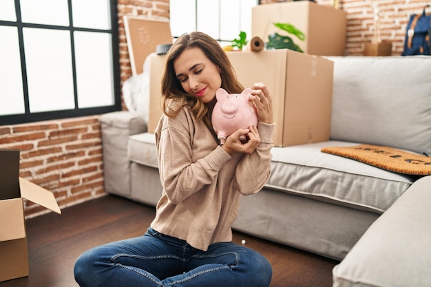 Young woman smiling confident hugging piggy bank at new home