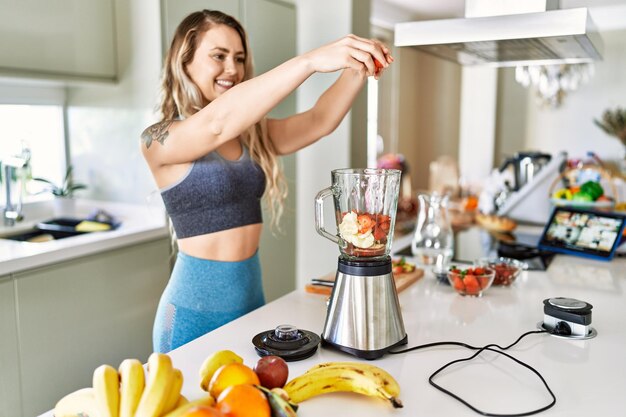 Young woman smiling confident pouring strawberries on blender at kitchen